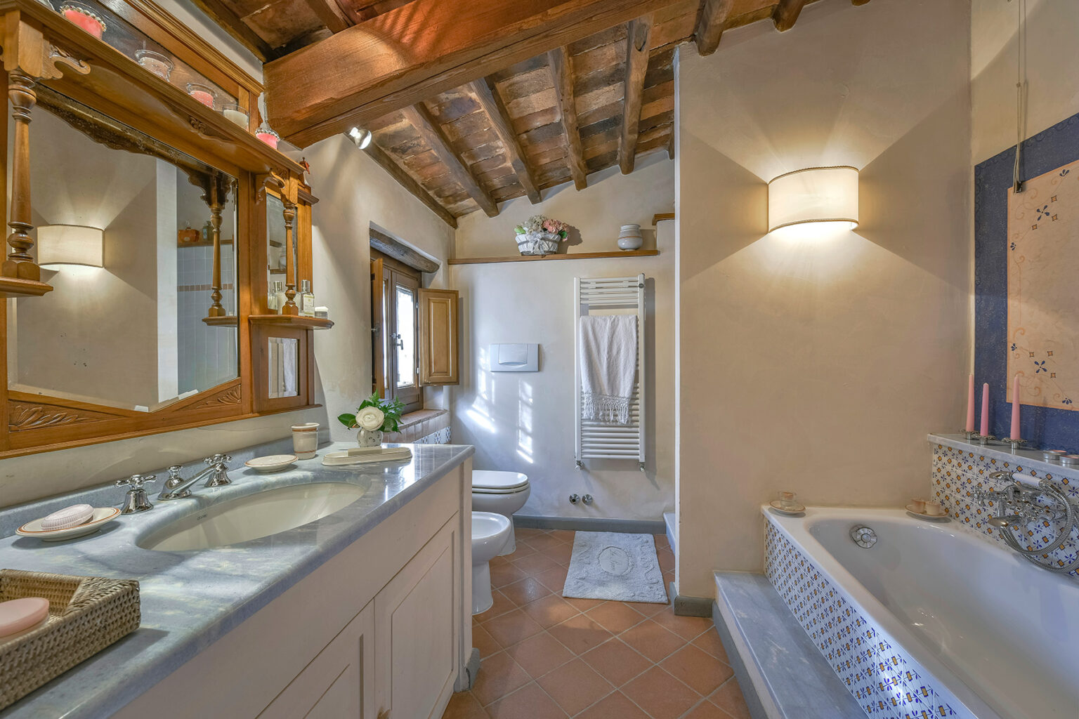Spacious bathroom featuring a double sink vanity, wooden ceiling beams, a large bathtub, and natural light from a window, creating a warm and inviting atmosphere. | Only on SopranoVillas