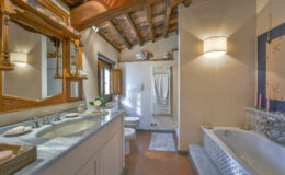 Spacious bathroom featuring a double sink vanity, wooden ceiling beams, a large bathtub, and natural light from a window, creating a warm and inviting atmosphere.