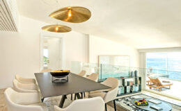 Modern dining area featuring a sleek black table surrounded by white chairs, complemented by stylish gold pendant lights. Large windows offer a stunning ocean view, enhancing the bright and airy atmosphere.