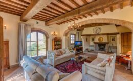 Cozy living room in a rustic home featuring wooden beams, a fireplace, comfortable sofas, and large windows with scenic views. Decorated with a chandelier and a wall clock, this inviting space combines traditional charm with modern amenities.