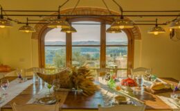 Rustic dining table set with elegant glassware and fresh produce, featuring large windows that offer a scenic view of the landscape. Warm lighting enhances the inviting atmosphere of the space.