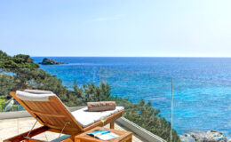 Lounge chair on a balcony overlooking a clear blue ocean with lush greenery in the background, perfect for relaxation and enjoying scenic coastal views.
