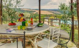 Outdoor dining area with a stone table set with colorful fruits and drinks, surrounded by olive trees and overlooking a scenic landscape with rolling hills and a pool in the background. Ideal for al fresco dining and relaxation.