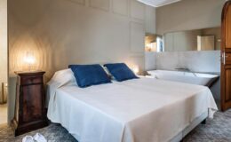 Cozy bedroom featuring a double bed with blue accent pillows, elegant wooden nightstand, and soft lighting, complemented by a modern bathroom area in the background. Ideal for relaxation and comfort.