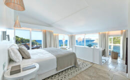 Spacious and modern bedroom with large windows offering ocean views, featuring a cozy bed with neutral bedding, a decorative rug, and stylish light fixtures, ideal for a relaxing getaway.