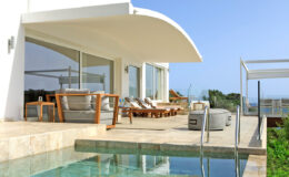 Luxury modern villa with a spacious outdoor terrace, comfortable lounge chairs, and an inviting swimming pool, overlooking a scenic view. Perfect for relaxation and entertainment in a sunny environment.