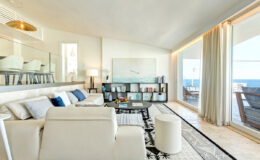 Modern coastal living room with white furniture, large windows offering ocean views, and a stylish bookshelf. Natural light floods the space, enhancing the airy atmosphere and elegant decor.