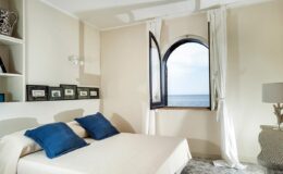 Cozy bedroom with a double bed featuring blue accent pillows, large window with ocean view, and minimalist decor, ideal for a relaxing getaway.