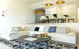 Modern living room featuring a spacious beige sofa with decorative pillows, a round coffee table with a fruit arrangement, and a stylish black and white rug. The background showcases a dining area with light-colored chairs and elegant lighting fixtures, creating a bright and inviting atmosphere.