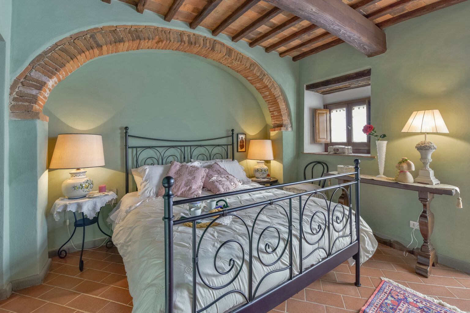Cozy bedroom featuring an iron bed frame, soft bedding, and warm lighting from bedside lamps, set against a rustic green wall with exposed wooden beams and a charming arched alcove. | Only on SopranoVillas