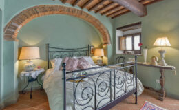 Cozy bedroom featuring an iron bed frame, soft bedding, and warm lighting from bedside lamps, set against a rustic green wall with exposed wooden beams and a charming arched alcove.
