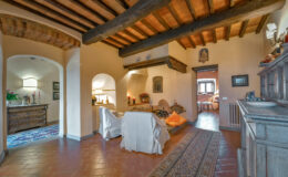 Cozy living room interior featuring rustic wooden beams, terracotta flooring, and warm lighting. The space includes comfortable seating, decorative art, and an inviting atmosphere, ideal for relaxation.