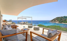 Luxurious outdoor seating area with wooden furniture overlooking a stunning coastal view, featuring clear blue waters and lush greenery. Ideal for relaxation and enjoying scenic vistas.