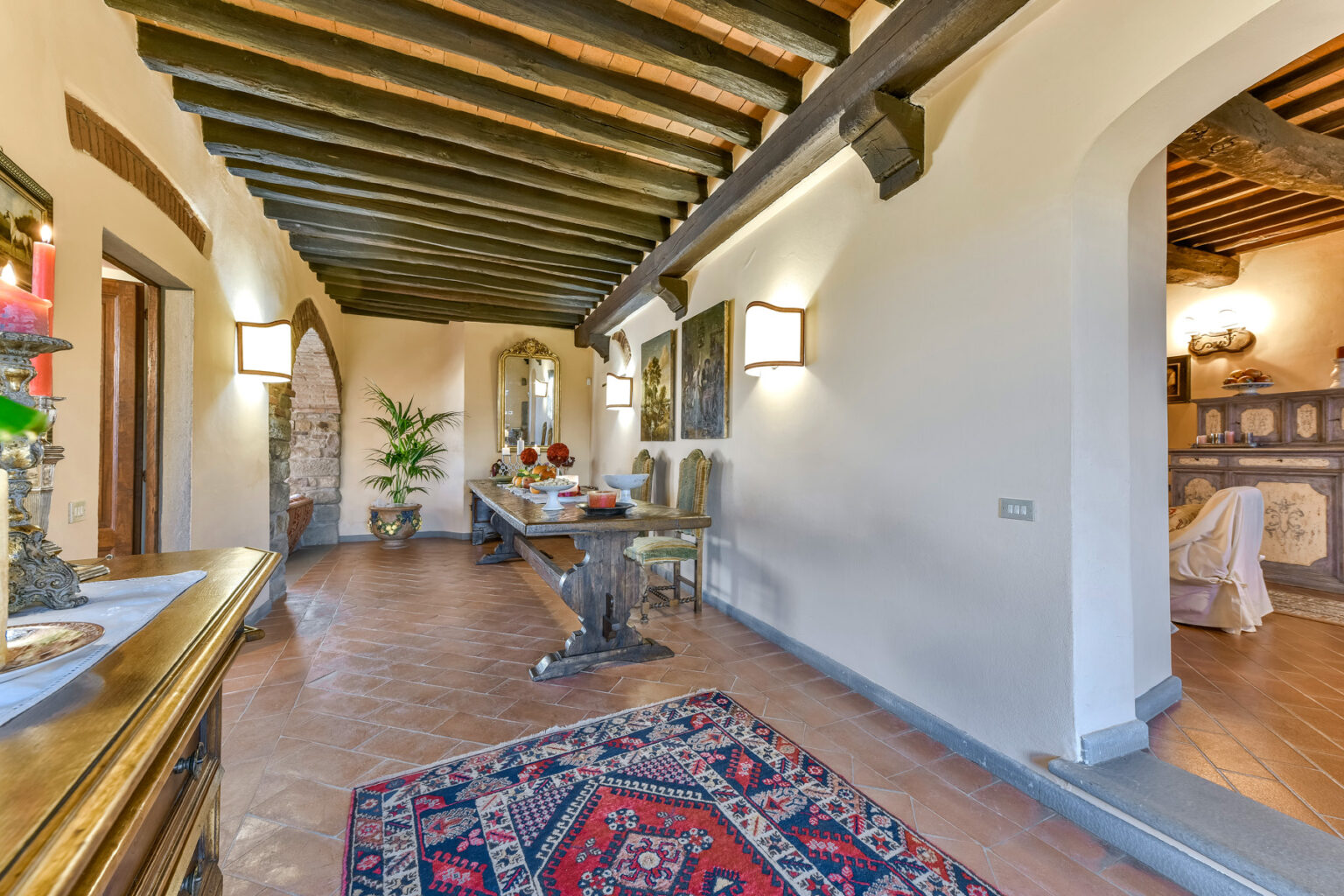 Elegant hallway with wooden beams, featuring a decorative rug, a side table with candles, and potted plants, leading to a cozy living area. | Only on SopranoVillas