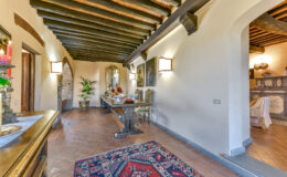 Elegant hallway with wooden beams, featuring a decorative rug, a side table with candles, and potted plants, leading to a cozy living area.