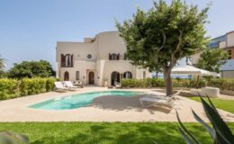Elegant villa with a private pool, surrounded by lush greenery and sun loungers, set against a clear blue sky. Perfect for a relaxing getaway.