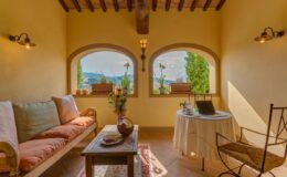 Cozy interior of a sunlit room featuring a comfortable sofa with cushions, a wooden coffee table, and a small dining table with a chair. Large windows offer scenic views of greenery, enhancing the inviting atmosphere. Ideal for relaxation or remote work.