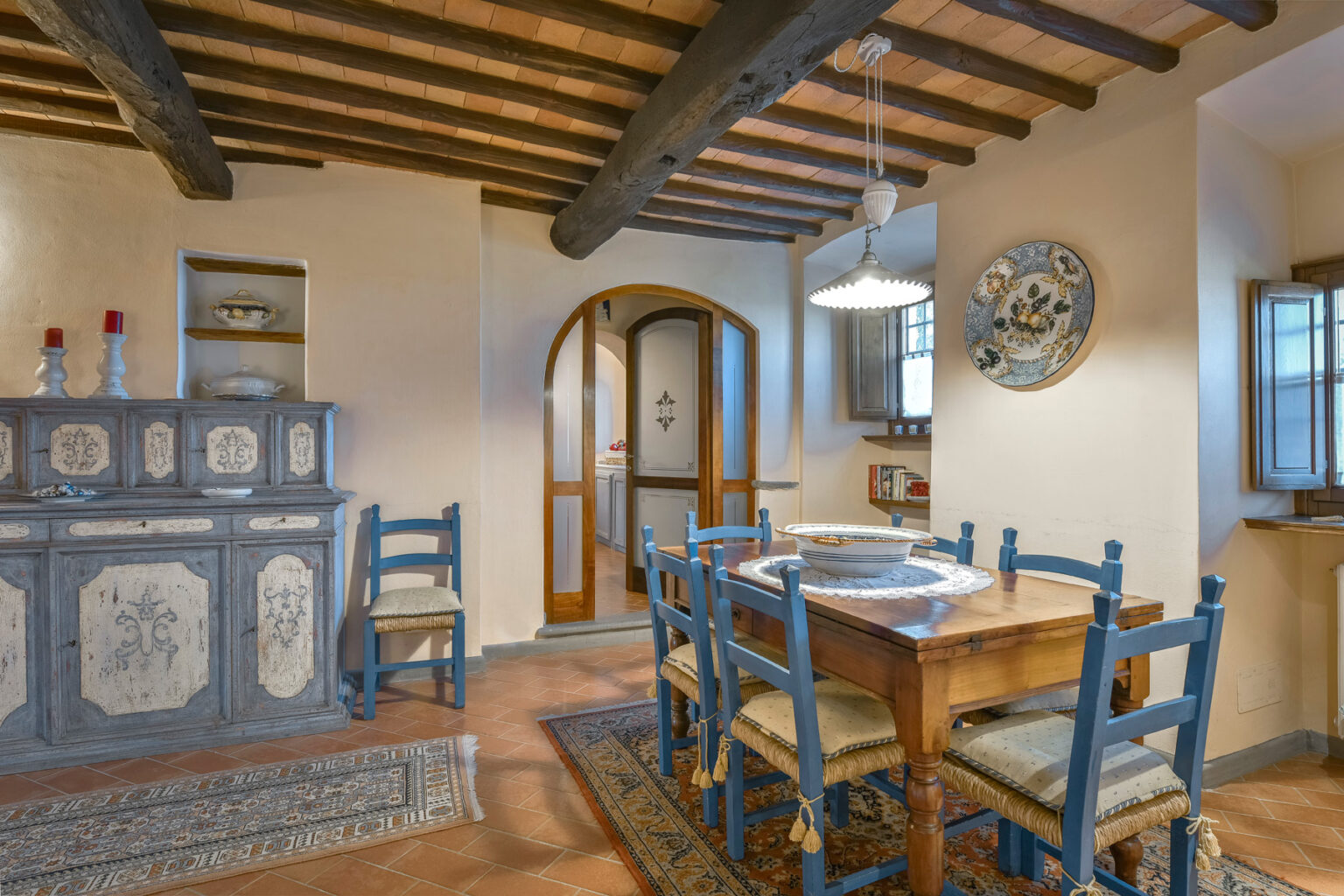 Cozy rustic kitchen interior featuring wooden beams, a dining table with blue chairs, and a decorative side cabinet, creating a warm and inviting atmosphere. | Only on SopranoVillas