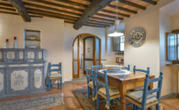 Cozy rustic kitchen interior featuring wooden beams, a dining table with blue chairs, and a decorative side cabinet, creating a warm and inviting atmosphere.