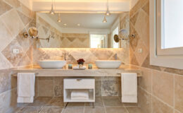 Modern bathroom interior featuring a double sink vanity with stylish bowls, elegant stone tile walls, and a well-lit mirror. Soft lighting enhances the sophisticated design, complemented by neatly arranged towels and decorative elements.