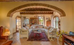 Cozy living room with a rustic design featuring a large beige sofa, decorative pillows, an ornate chandelier, and a warm color palette. Natural light streams through arched windows, illuminating a traditional area rug and wooden furniture. Ideal for showcasing comfortable and inviting home interiors.