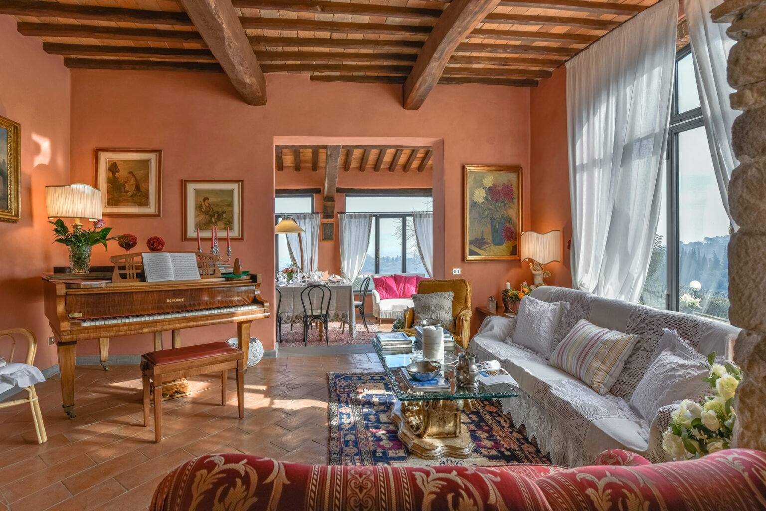 Cozy living room featuring wooden beams, warm peach walls, a grand piano, elegant furniture, and natural light streaming through large windows, creating an inviting atmosphere. | Only on SopranoVillas