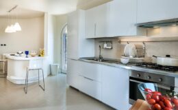 Modern kitchen interior featuring sleek white cabinetry, a gas stove, and a marble countertop, complemented by a dining area with bar stools and natural light streaming in from a nearby window.