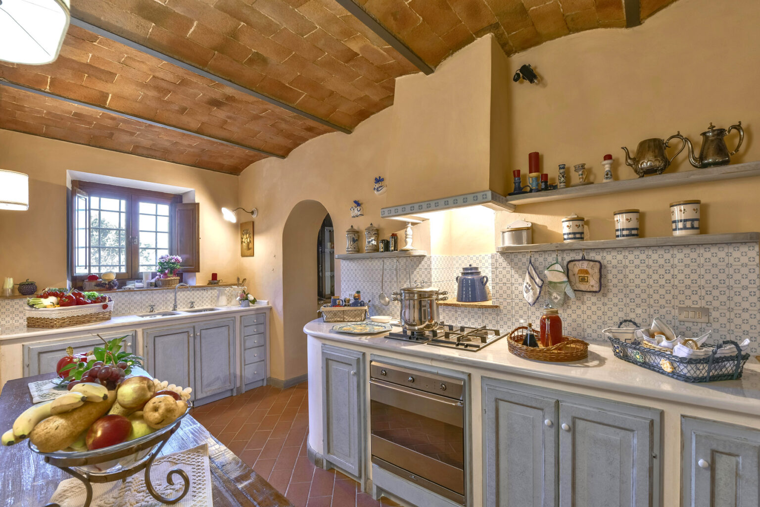 Rustic kitchen interior with wooden ceiling, light-colored cabinetry, and modern appliances. Features a large window, decorative shelves with kitchenware, and a fruit bowl on the table, creating a warm and inviting atmosphere. Ideal for home design inspiration. | Only on SopranoVillas