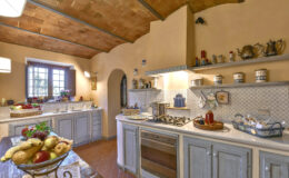 Rustic kitchen interior with wooden ceiling, light-colored cabinetry, and modern appliances. Features a large window, decorative shelves with kitchenware, and a fruit bowl on the table, creating a warm and inviting atmosphere. Ideal for home design inspiration.