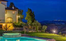 Scenic view of a luxury villa with a swimming pool at dusk, surrounded by lush greenery and illuminated by outdoor lighting, showcasing the serene landscape of rolling hills in the background.