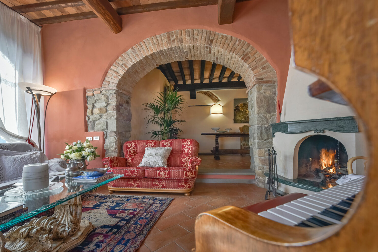Cozy living room interior featuring a red vintage sofa, glass coffee table, and a piano, with a stone archway leading to a dining area and a fireplace, complemented by warm lighting and decorative plants. | Only on SopranoVillas
