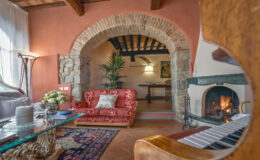 Cozy living room interior featuring a red vintage sofa, glass coffee table, and a piano, with a stone archway leading to a dining area and a fireplace, complemented by warm lighting and decorative plants.
