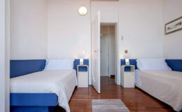 Cozy bedroom featuring two single beds with blue accents, bedside tables, and a bright, airy atmosphere. The room has wooden flooring and a door leading to another area, ideal for a comfortable stay.