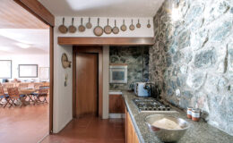 Modern kitchen with stone walls, featuring wooden utensils hanging, a granite countertop, and a bowl of dough. The space connects to a dining area with wooden furniture, creating a cozy atmosphere.