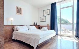 Modern bedroom with a large window, wooden flooring, and minimalist decor, featuring a double bed with white linens, a bedside lamp, and artwork on the walls. Sunlight streams in, highlighting the spacious and inviting atmosphere.