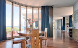 Modern dining area with a round wooden table and chairs, featuring large windows with blue curtains that offer a scenic view. The space has hardwood flooring and is connected to an open kitchen area, creating a bright and inviting atmosphere.