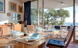 Spacious dining area with a wooden table set for a meal, featuring blue dishware and wine, surrounded by large windows showcasing a scenic view of the ocean and greenery.
