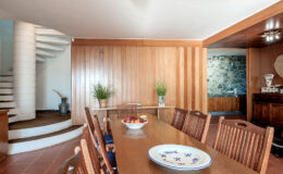 Modern dining room featuring a large wooden table surrounded by chairs, with a spiral staircase in the background and decorative plants. The warm wood paneling adds a cozy atmosphere, complemented by a stylish plate centerpiece.