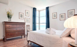 Cozy bedroom featuring a comfortable bed with white linens, a wooden dresser, and large windows with blue curtains, allowing natural light to illuminate the space. Decorated with framed artwork and a stylish lamp, this inviting room offers a serene atmosphere.