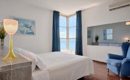 Cozy bedroom with a white bed, blue curtains, and a scenic ocean view, featuring a decorative yellow glass lamp and a blue armchair.