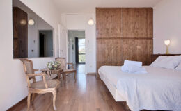Modern bedroom with wooden accents, featuring a cozy bed, minimalist decor, and natural light streaming through large windows. A small seating area with two chairs and a table adds charm to the space.
