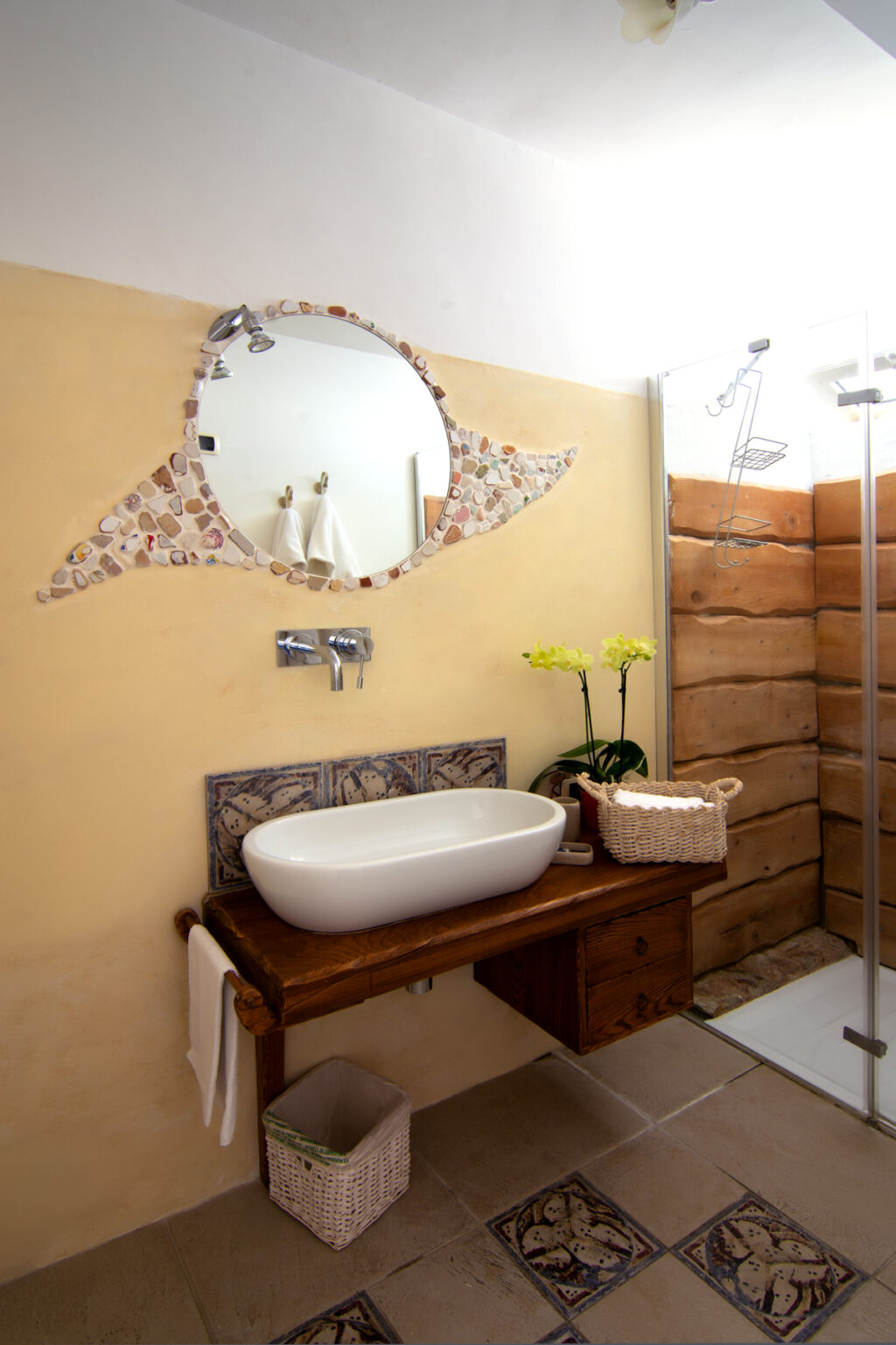 Modern bathroom interior featuring a stylish white vessel sink on a wooden vanity, decorative mirror with mosaic design, and a potted orchid. Natural wood accents and warm wall color create a cozy atmosphere. | Only on SopranoVillas