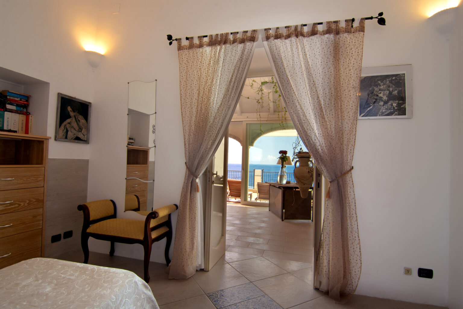 Cozy bedroom interior with a view of the sea, featuring sheer curtains, a wooden dresser, and a stylish chair, creating a relaxing atmosphere. | Only on SopranoVillas