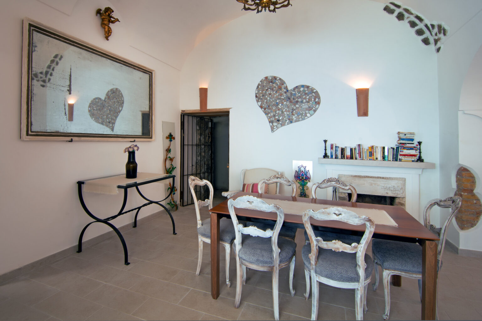 Cozy dining area featuring a wooden table with elegant chairs, decorative heart-shaped wall art, and a large mirror. Soft lighting enhances the inviting atmosphere, making it a perfect space for gatherings. | Only on SopranoVillas
