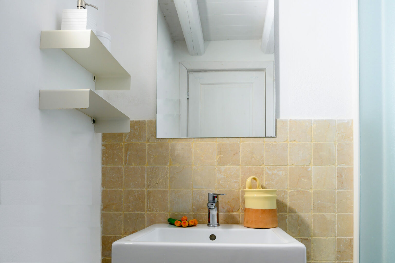 Modern bathroom with a white sink, yellow accents, and a mirror, featuring a tiled wall and minimalist shelving. Fresh fruits are placed next to the sink, enhancing the clean and inviting atmosphere. | Only on SopranoVillas