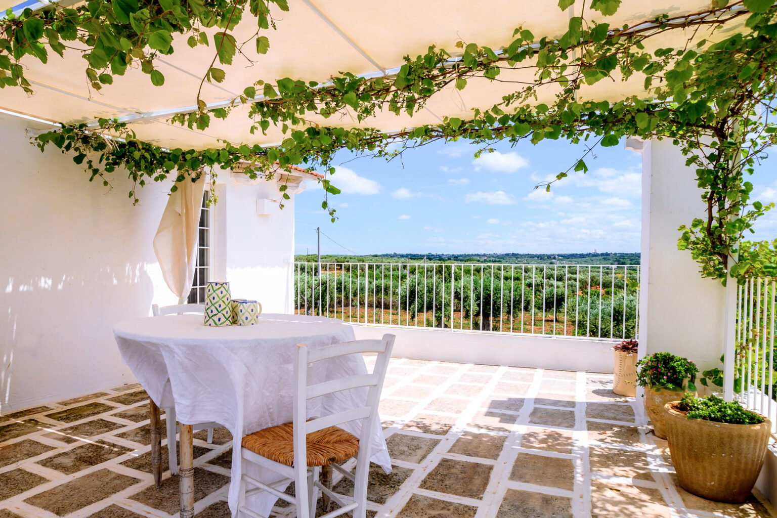 Cozy outdoor dining area with a white table and chairs, surrounded by lush greenery and a scenic view of olive groves under a bright blue sky. Ideal for relaxation and al fresco meals. | Only on SopranoVillas