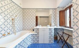 Bright and airy bathroom featuring intricate blue and white patterned tiles, a spacious bathtub, a window with natural light, and a stylish wooden folding chair.