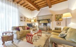 Cozy living room featuring a wooden beam ceiling, comfortable sofas with decorative pillows, a traditional fireplace, and framed artwork on the walls, illuminated by natural light from large windows.