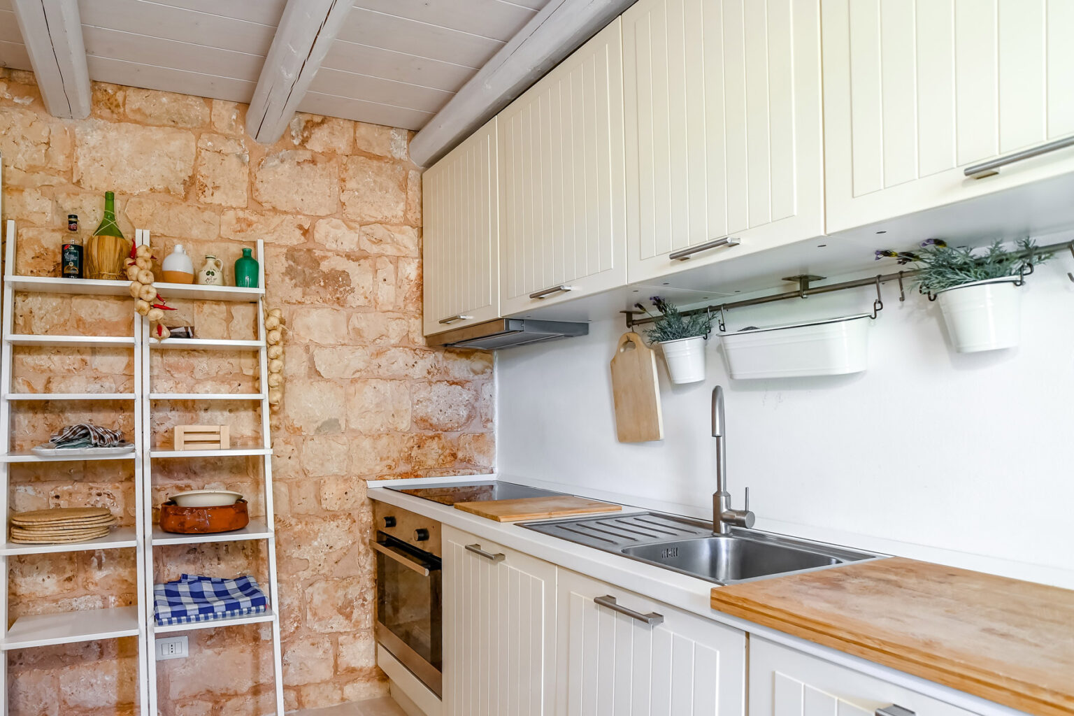 Modern kitchen with white cabinetry, wooden countertops, and a stone accent wall. Features a stainless steel sink, an oven, and open shelving with decorative items and kitchen essentials. Bright and airy atmosphere with natural light. | Only on SopranoVillas