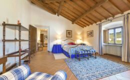 Spacious bedroom with wooden beam ceiling, featuring a large bed with blue and white bedding, a cozy seating area, and a patterned area rug. Natural light streams in through the window, illuminating the rustic decor and warm tones of the room.